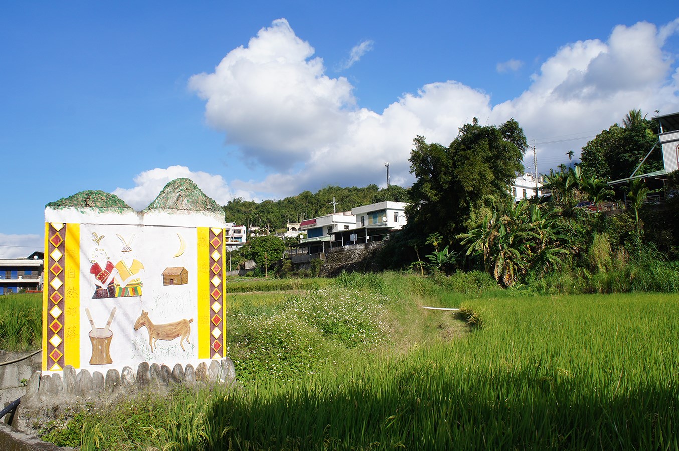 滿自然部落入口彩繪意象,畫面包含人物、山羌圖案,後方可見稻田與聚落